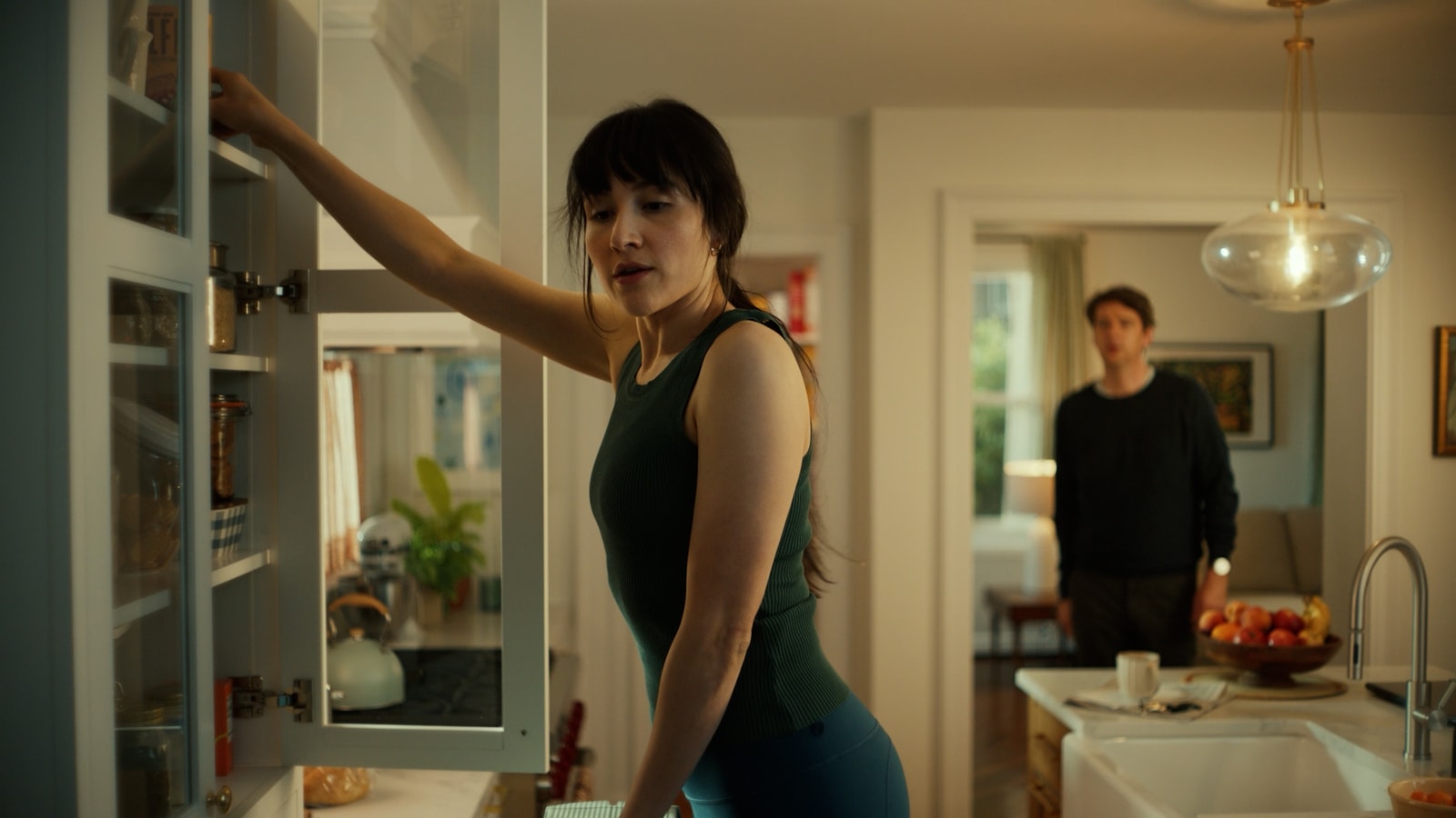 A woman reaches into a cupboard while a man stands behind her in a kitchen.