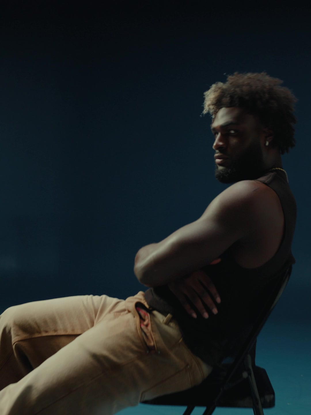 Will Anderson Jr. sits in a black sleeveless top and tan trousers against a dark blue studio background.