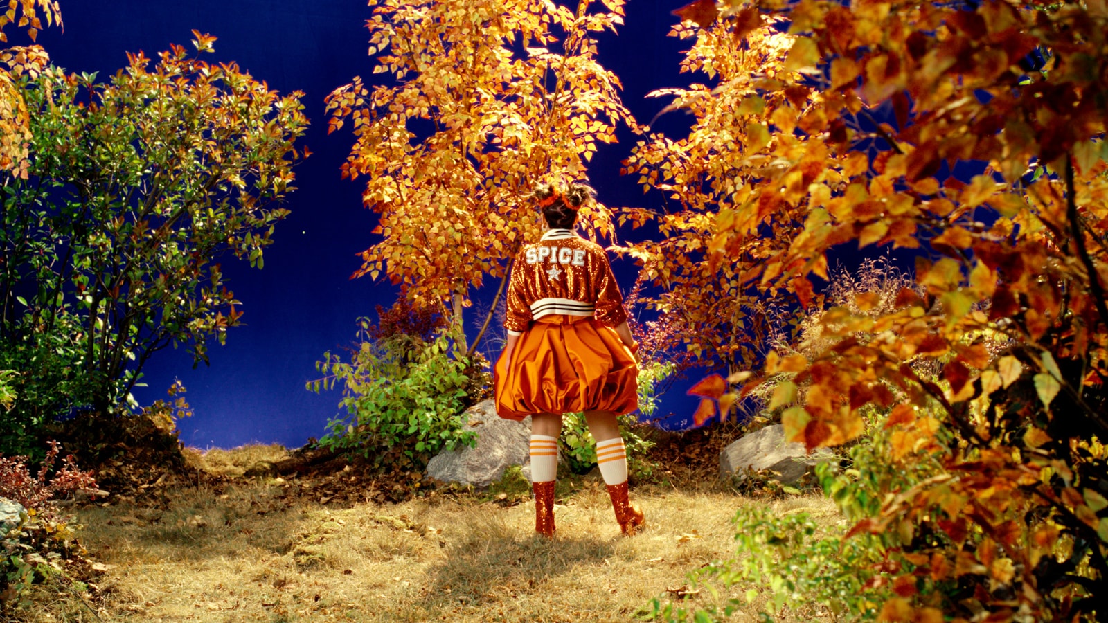 Rachel Dratch stands among practical autumn trees and rocks in front of a blue screen on the music-video set.