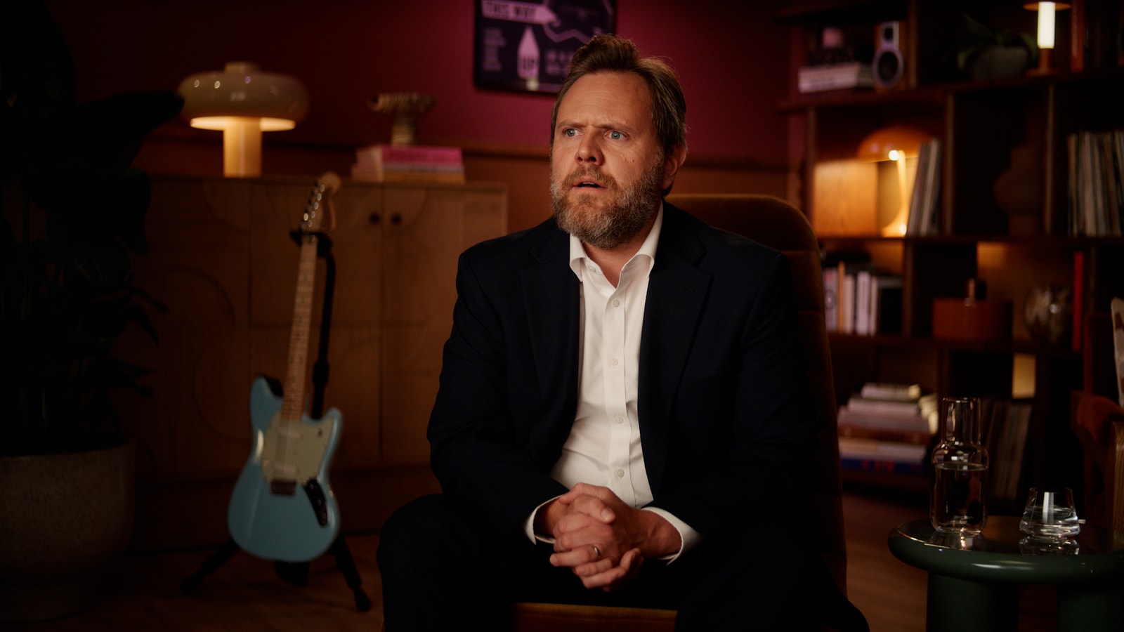 An interviewer in a dark suit sits in the recording studio during the Pumpkin Spice reunion.