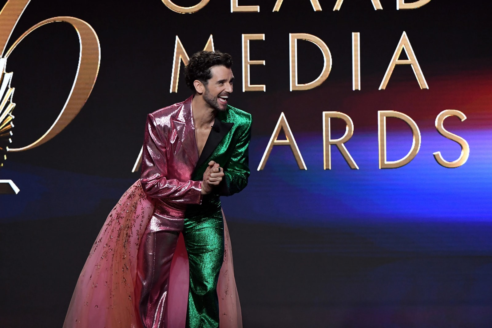 A performer in a red and green suit holds a microphone on the 2025 GLAAD stage.