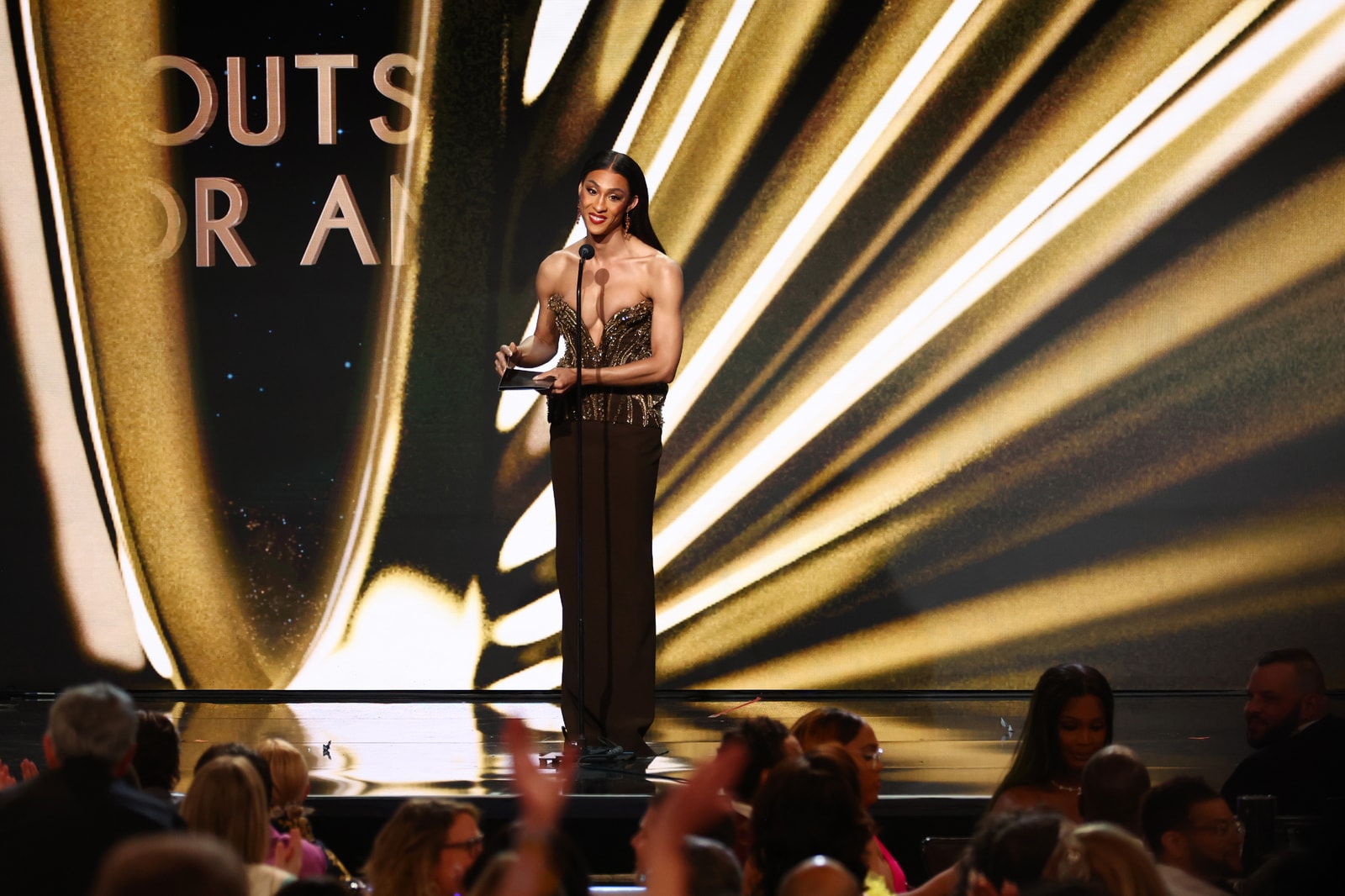 A presenter at a podium against radiating gold statuette graphics.