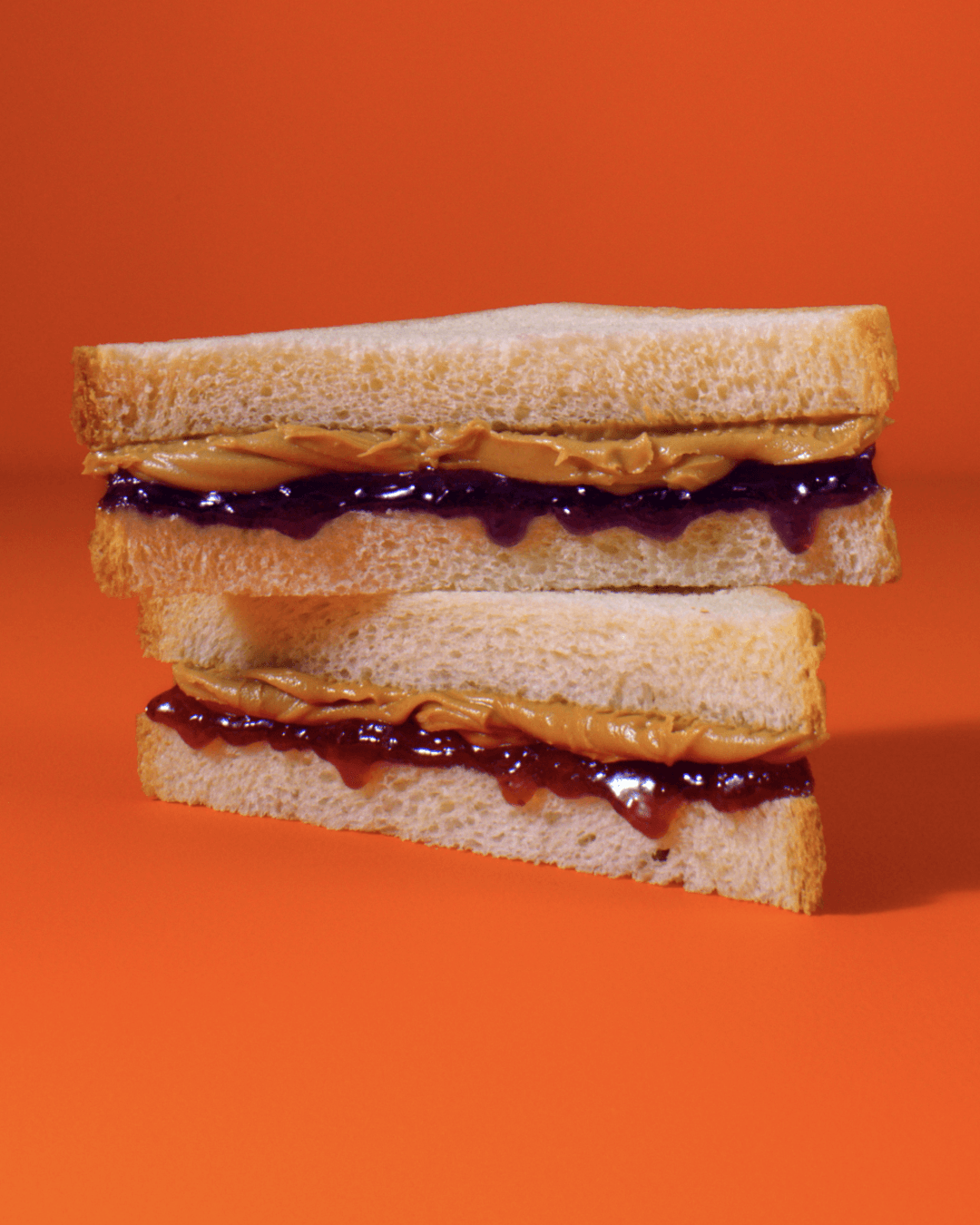 Two peanut butter and jelly sandwich halves stacked on an orange background.