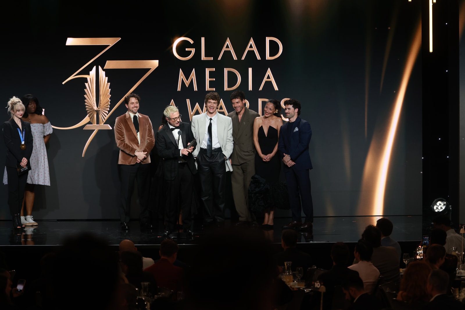 A group stands before the gold 37th GLAAD Media Awards stage graphic.