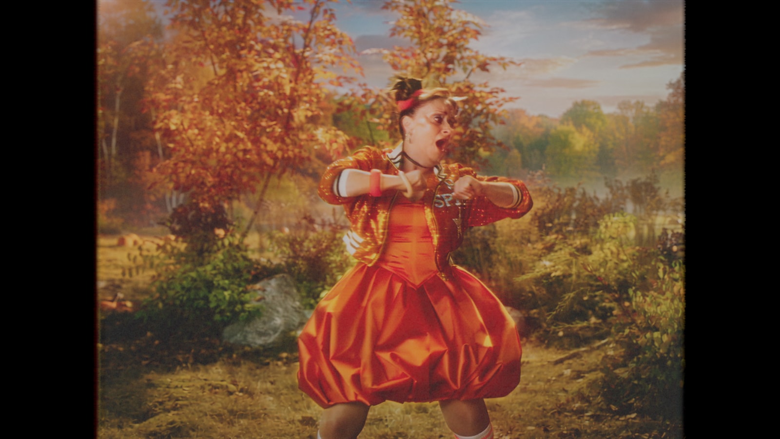Rachel Dratch performs a dance move in an orange pumpkin dress against the music video’s autumn woodland background.