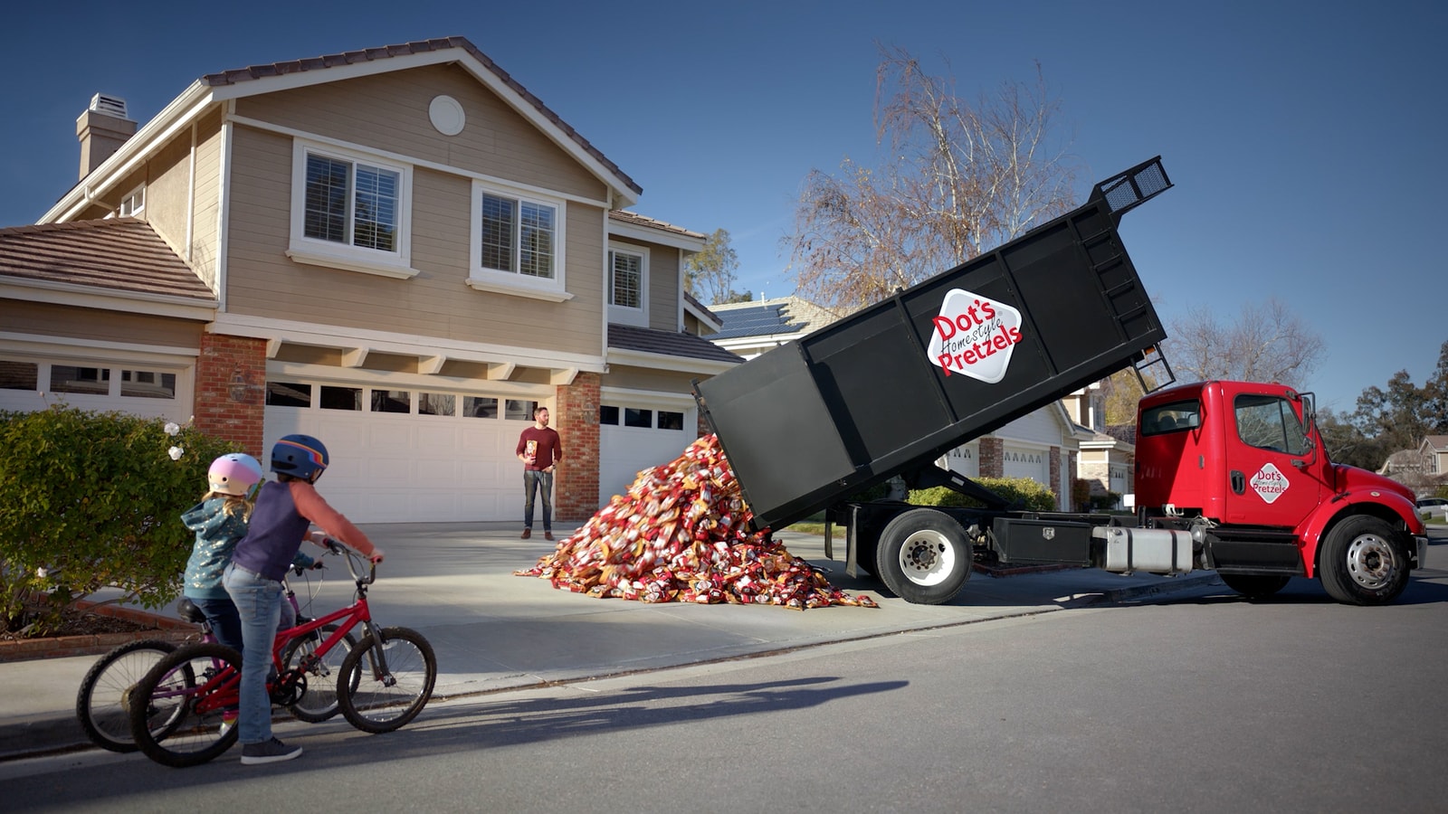 Cyclists pass a dump truck unloading pretzel bags outside a house.