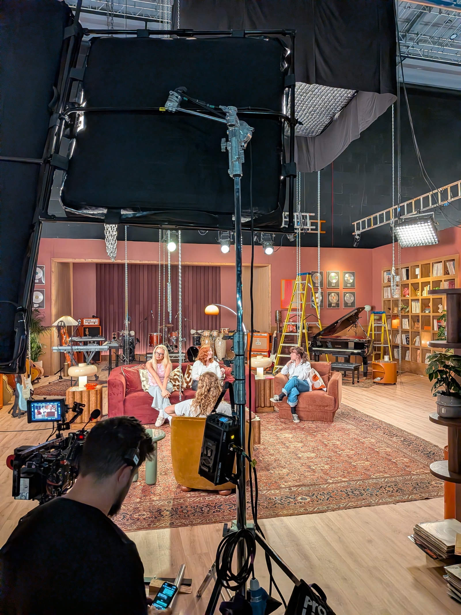 A crew member and overhead lighting equipment frame the recording-studio set during filming.