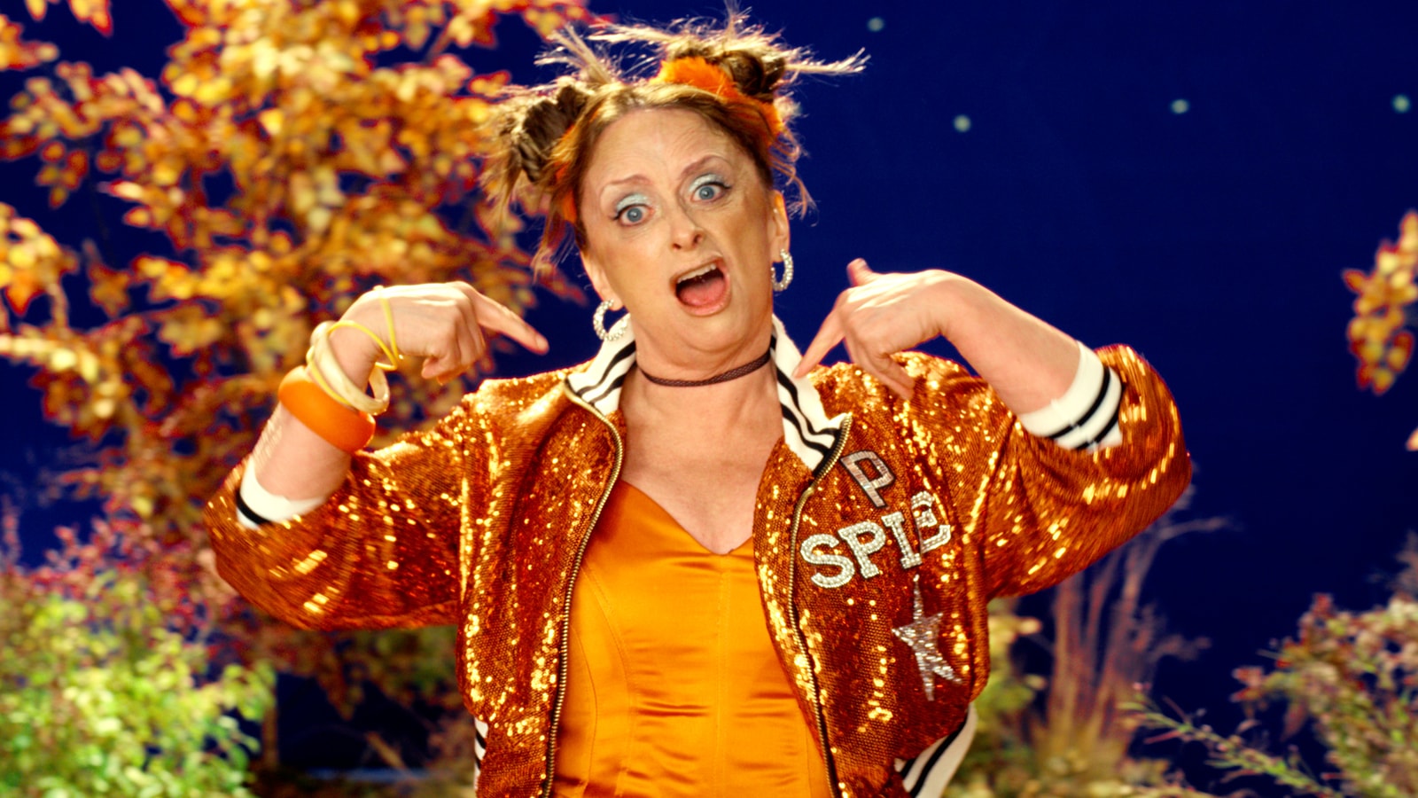 Rachel Dratch points toward her sequined Pumpkin Spice jacket while performing in front of the blue screen.